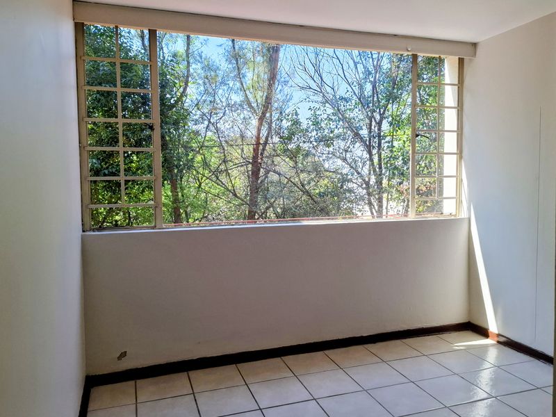 A bright, empty bedroom featuring tiled flooring and a large window with security bars, offering a pleasant view of mature trees.
