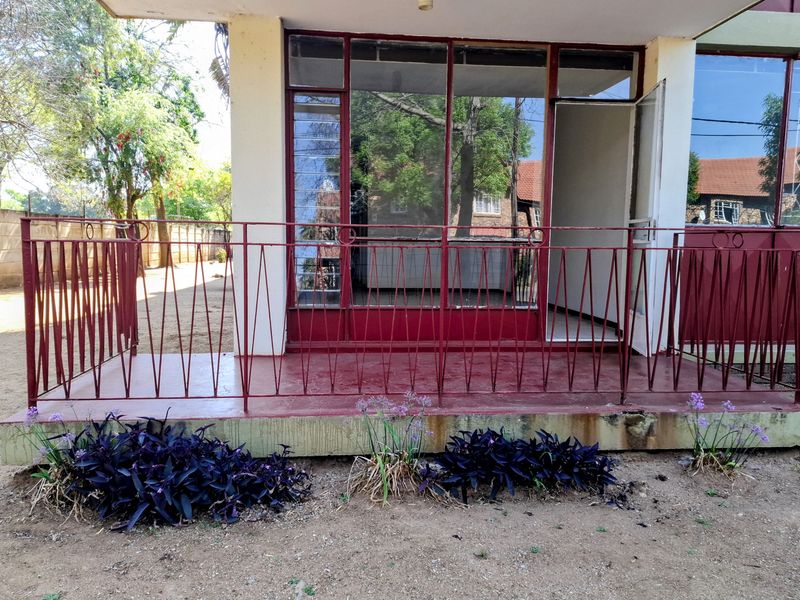 A ground-level patio area featuring a red metal balustrade, concrete flooring, and aluminium-framed glass doors leading into the apartment.