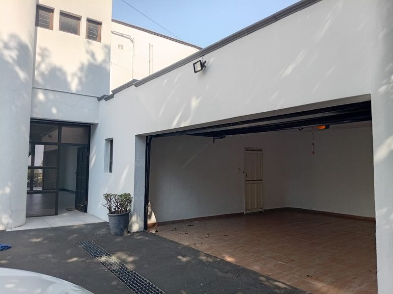 A view of an open-plan double garage with tiled flooring, adjacent to the main building entrance and paved driveway area.