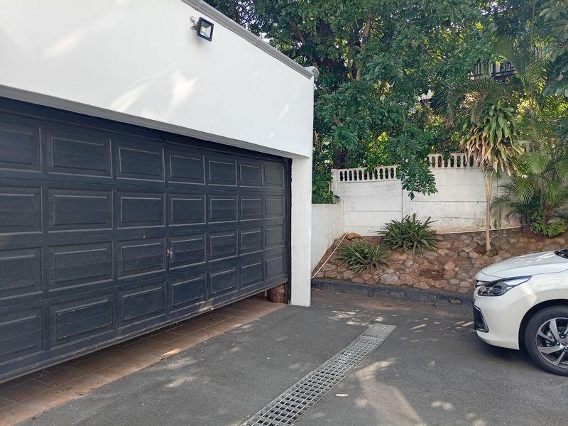 Exterior view of a garage featuring a dark sectional door, paved driveway with drainage channel, and a landscaped retaining wall.