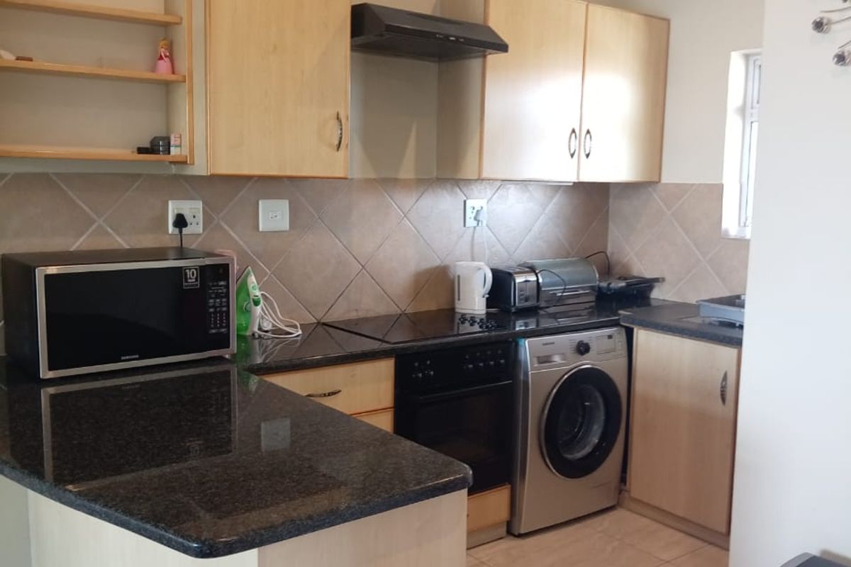 A functional kitchen featuring granite countertops, light wood cabinetry, a tiled backsplash, and built-in appliances including an oven, hob, and washing machine.