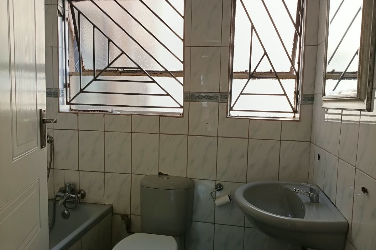 A dated bathroom featuring a pedestal basin, toilet, and bathtub with tiled walls and security-barred windows, requiring cosmetic updates and maintenance.