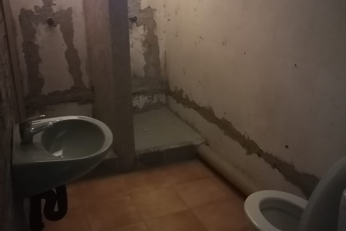 An unfinished bathroom space featuring a wall-mounted basin, toilet, and tiled floor, currently requiring significant renovation and wall finishing to be functional.