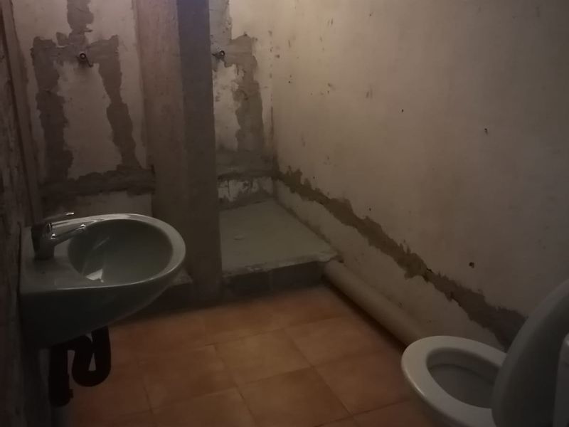 An unfinished bathroom space featuring a wall-mounted basin, toilet, and tiled floor, currently requiring significant renovation and wall finishing to be functional.