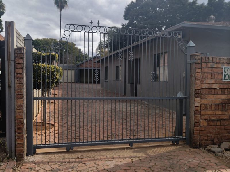 Front entrance view featuring a secure wrought iron sliding gate, brick boundary wall, and a paved driveway leading to a carport.
