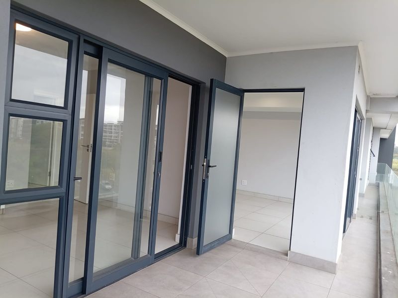 A modern, covered balcony featuring tiled flooring, sleek aluminium-framed sliding glass doors, and a contemporary glass balustrade overlooking the surrounding area.