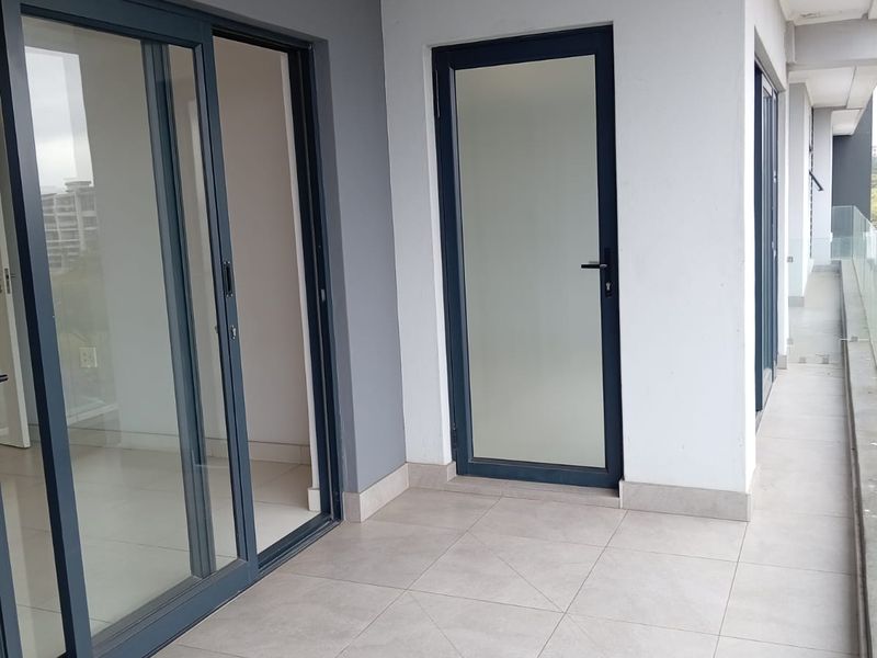 A modern, clean balcony featuring large sliding glass doors, a frosted glass entry door, and neutral floor tiles with a glass balustrade.