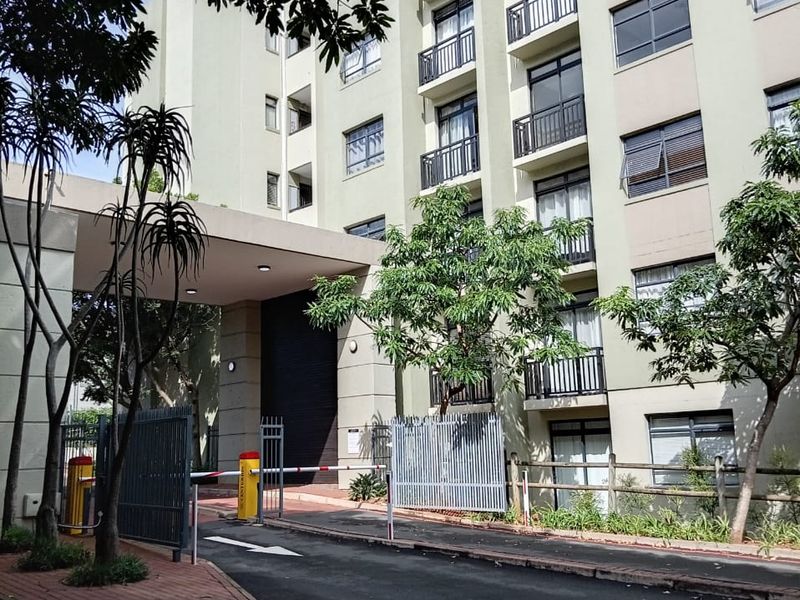 Exterior view of the apartment building entrance featuring a secure boom gate, paved driveway, and multi-story facade with private balconies.