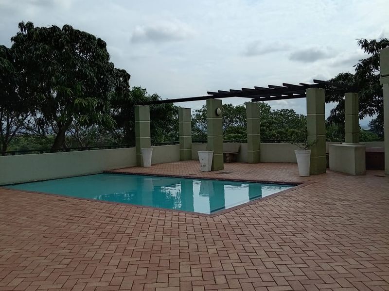 A spacious outdoor pool area featuring brick paving, a pergola structure, and concrete pillars, offering a clean and well-maintained recreational space.