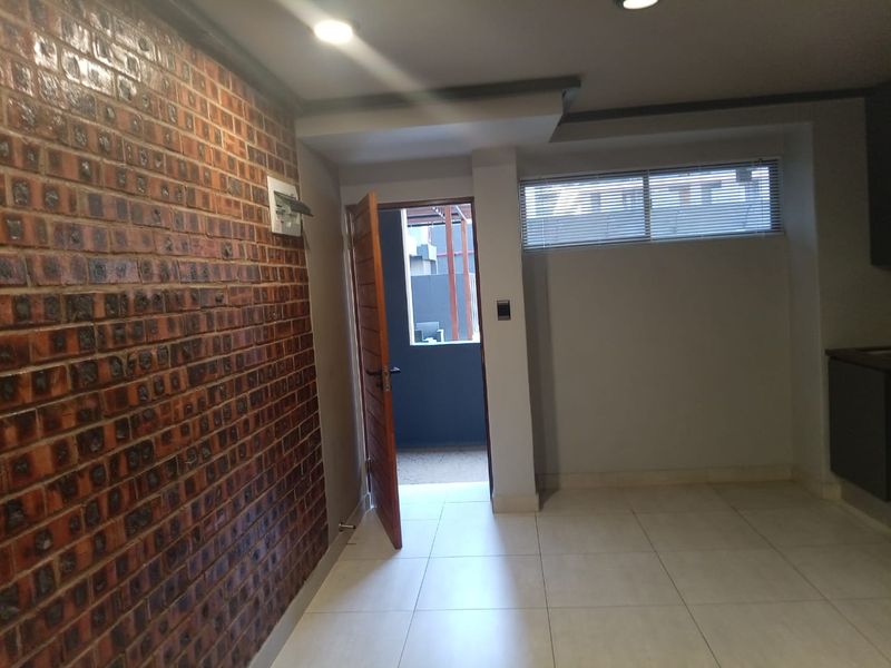 An open-plan living area featuring a distinctive exposed brick feature wall, neutral tiled flooring, recessed lighting, and direct access to an outdoor space.