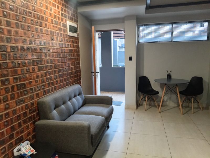 A living area featuring an exposed brick feature wall, neutral tiled flooring, and natural light through a window with venetian blinds.
