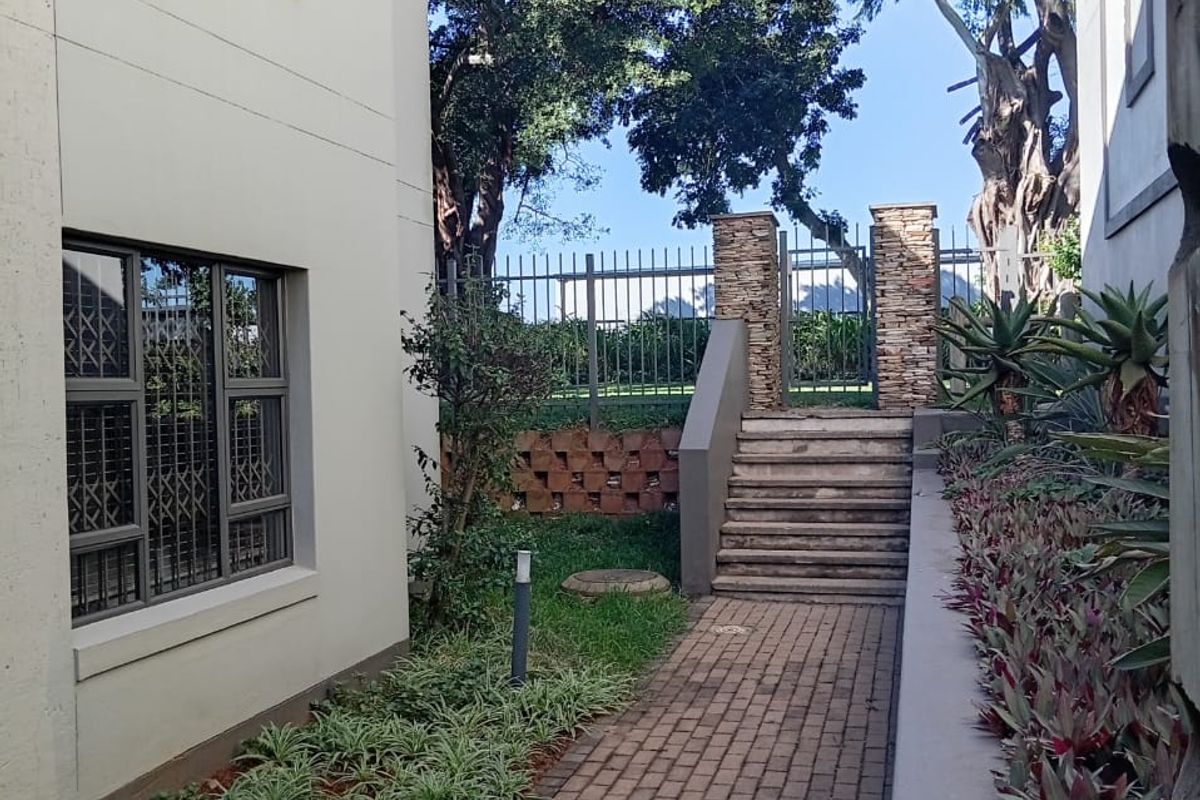 A paved walkway leads to a set of concrete stairs and a gated entrance, flanked by landscaped garden beds and the apartment building exterior.