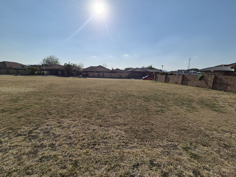 A wide-angle view of a vacant residential plot featuring dry grass and a surrounding brick boundary wall in a suburban neighborhood.