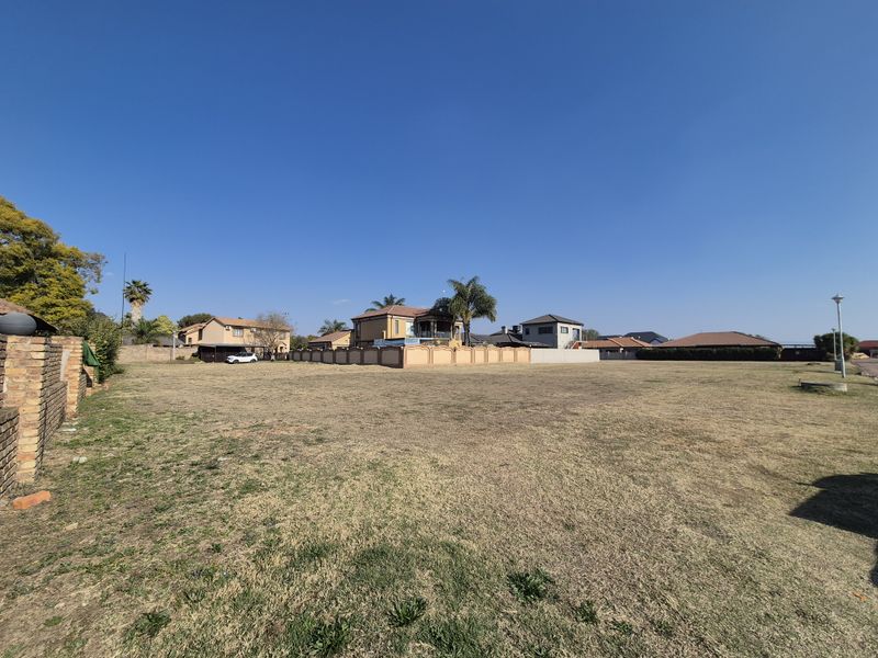 A wide-angle view of a vacant residential plot featuring dry grass, a perimeter boundary wall, and surrounding neighborhood homes.