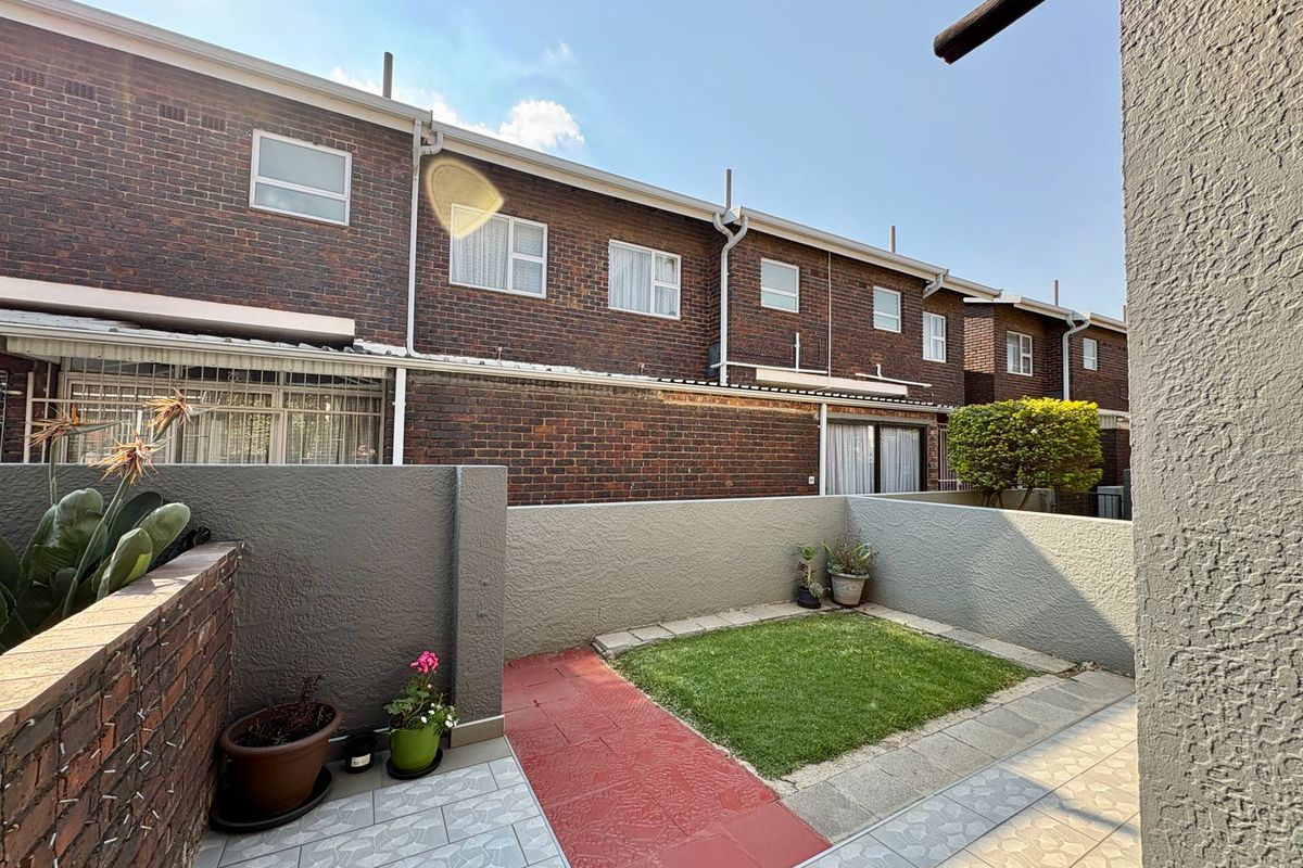 A private rear courtyard featuring a small lawn, paved walkway, and tiled patio area, enclosed by painted boundary walls against a brick townhouse facade.
