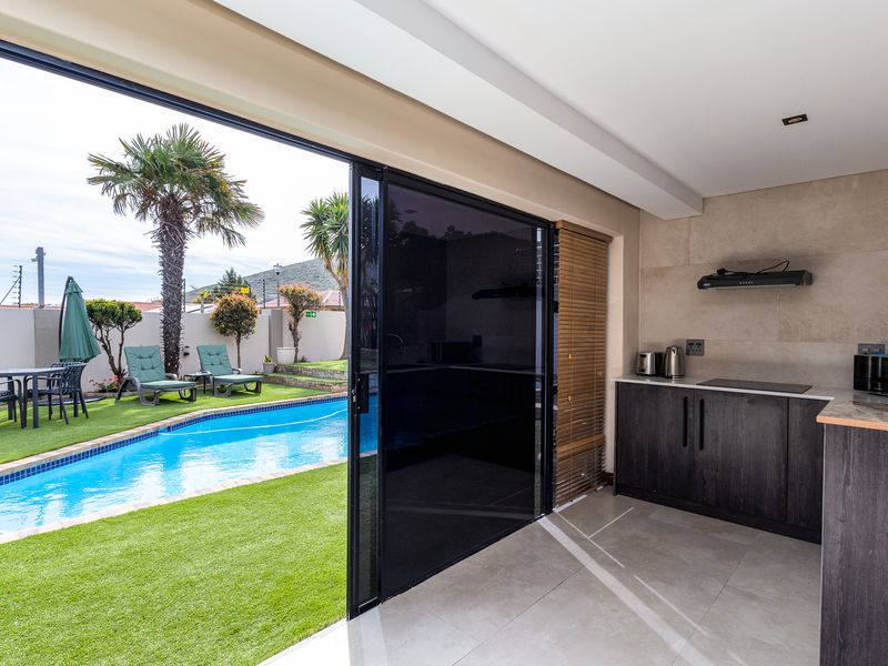 Modern open-plan indoor-outdoor living area featuring a kitchenette with built-in hob, flowing seamlessly onto a patio with a swimming pool and garden.