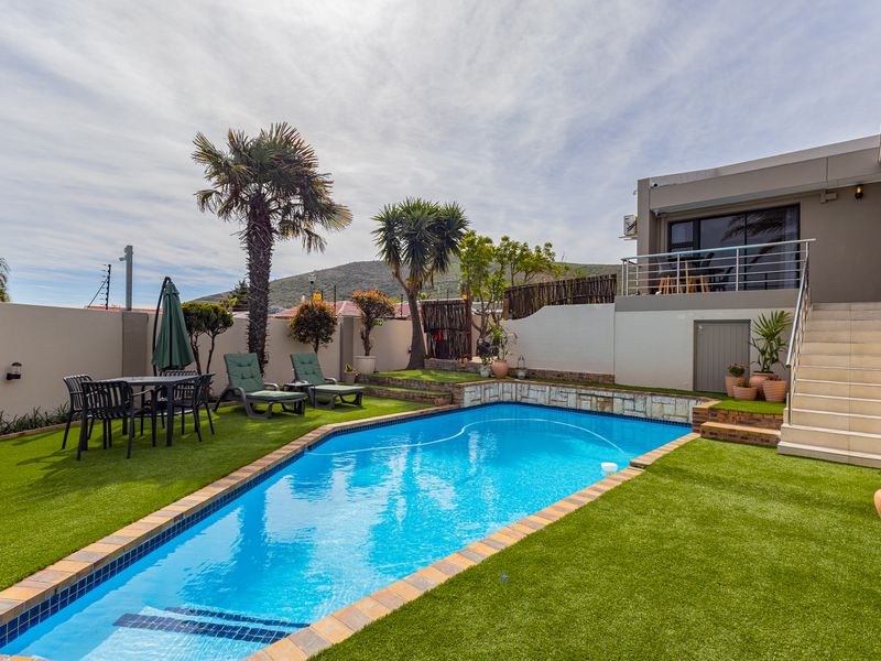 A well-maintained outdoor pool area featuring a swimming pool with stone coping, artificial grass, and a staircase leading to an elevated patio.