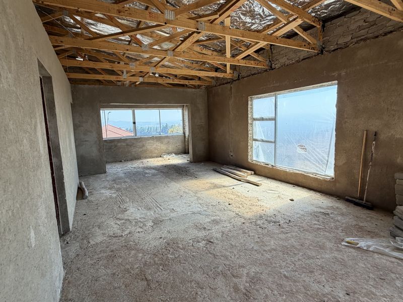 An unfinished living area featuring exposed timber roof trusses, cement screed flooring, and large aluminium windows with views, currently in a shell construction state.