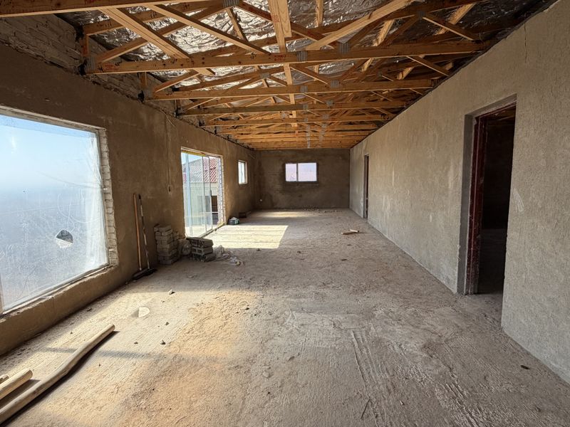 An unfinished open-plan interior space featuring exposed timber roof trusses, plastered walls, and large aluminium windows, currently in a shell construction state.