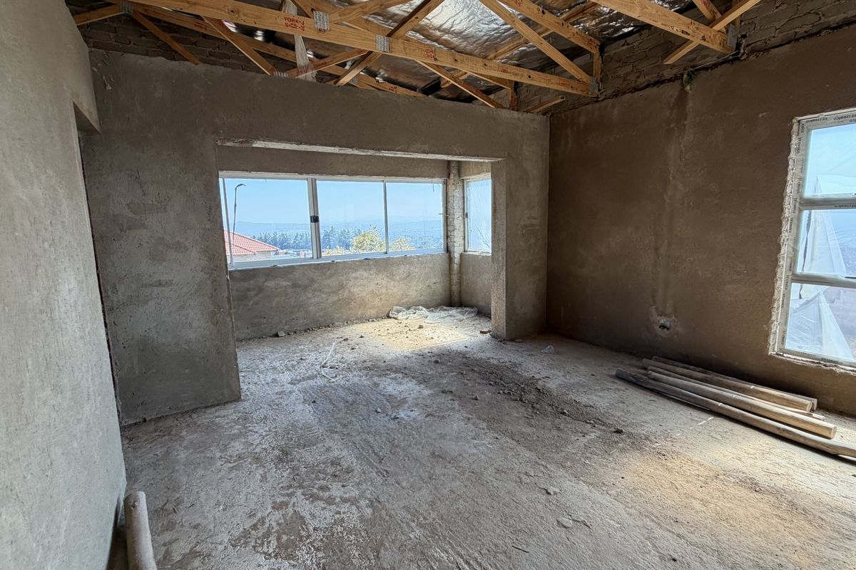 An unfinished bedroom interior in shell condition, featuring exposed timber roof trusses, cement plaster walls, and aluminium window frames with scenic views.