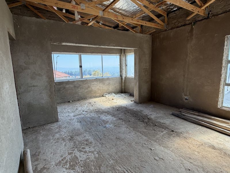 An unfinished bedroom interior in shell condition, featuring exposed timber roof trusses, cement plaster walls, and aluminium window frames with scenic views.