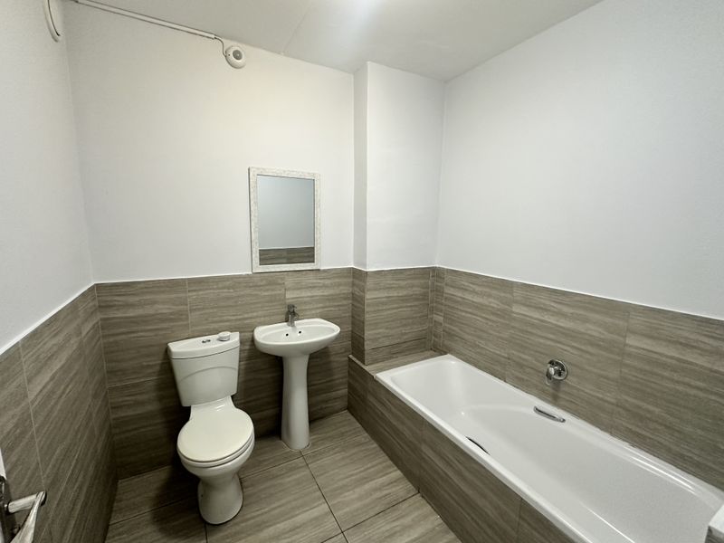 A clean, modern bathroom featuring a pedestal washbasin, toilet, and bathtub with neutral wood-look wall and floor tiling.