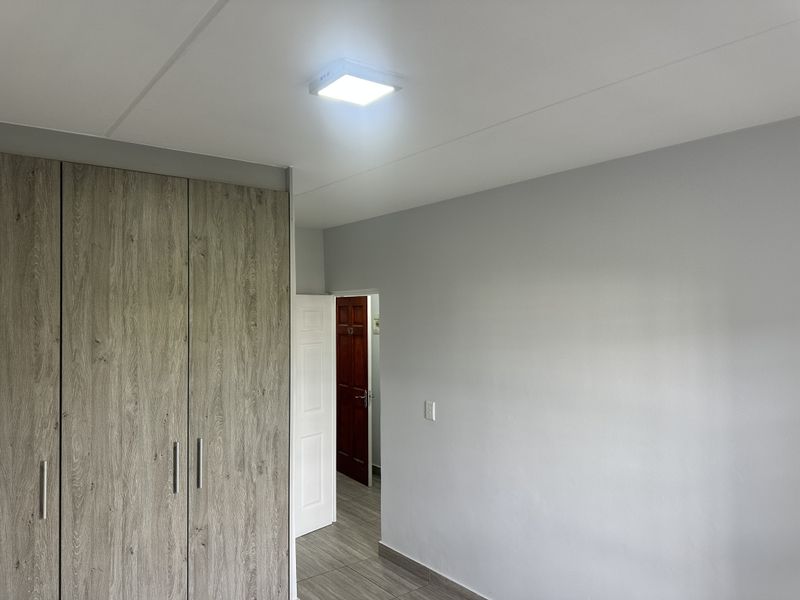 A clean, empty bedroom featuring modern wood-look built-in cupboards, neutral grey walls, and tiled flooring with a bright overhead light fixture.