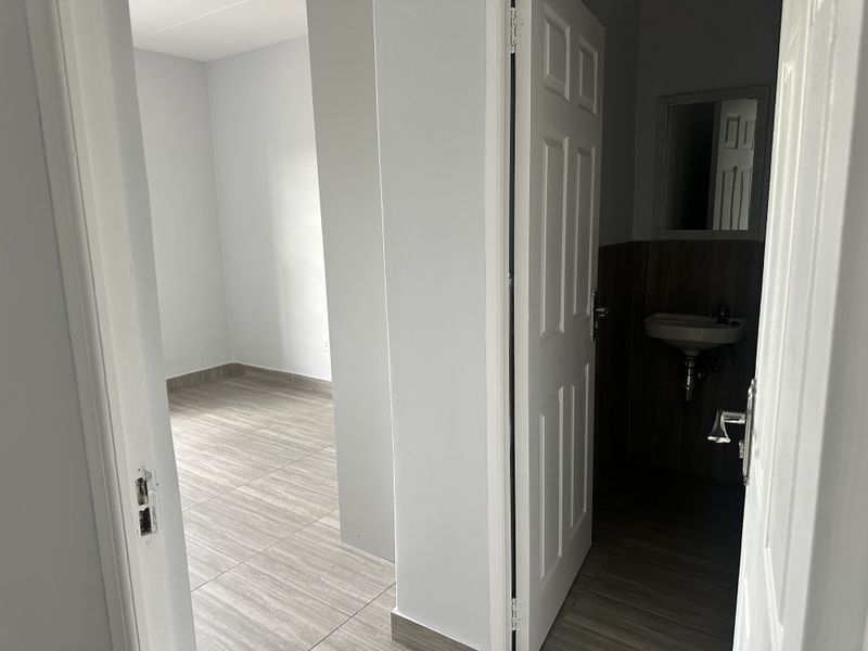 A view from a hallway looking into an empty bedroom and a small bathroom featuring a wall-mounted basin and neutral tiled flooring.