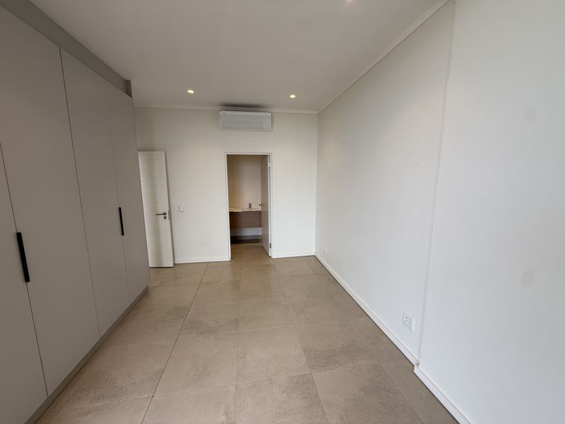 A clean, empty bedroom featuring neutral tiled flooring, built-in cupboards, and a wall-mounted air conditioning unit, with an adjoining en-suite bathroom.
