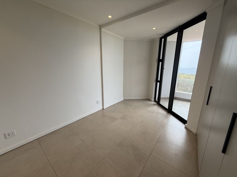 A bright, empty bedroom featuring neutral tiled flooring, built-in cupboards, and large sliding glass doors that open to a balcony with coastal views.