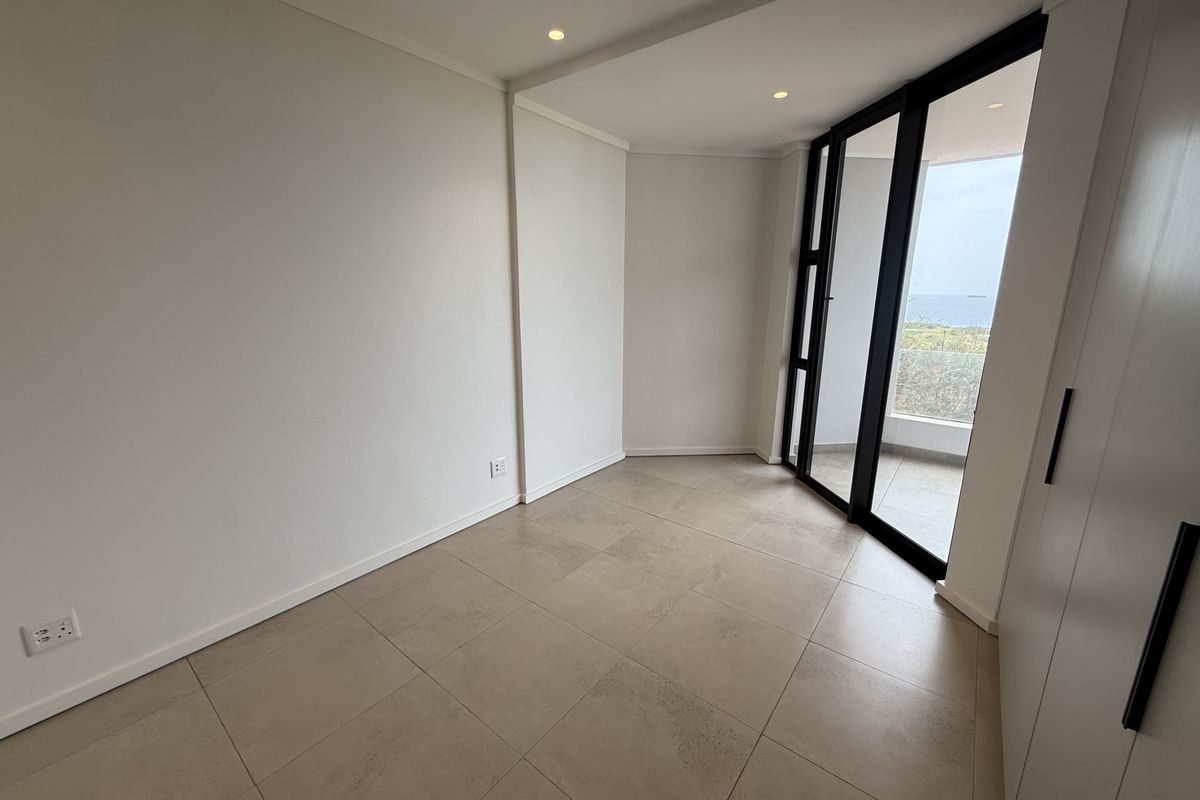 A bright, empty bedroom featuring neutral tiled flooring, built-in cupboards, and large sliding glass doors that open to a balcony with coastal views.