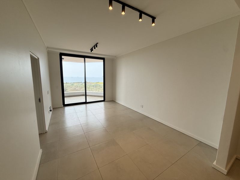 A bright, empty living area featuring large sliding glass doors that open to a balcony with scenic ocean views and modern track lighting.