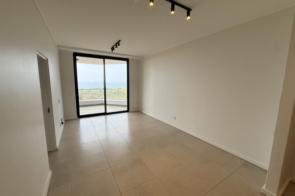 A bright, empty living area featuring large sliding glass doors that open to a balcony with scenic ocean views and modern track lighting.