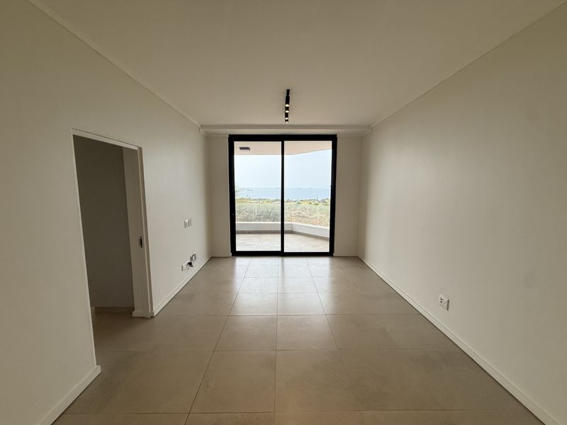 A bright, empty living area featuring large sliding glass doors that open to a balcony with scenic ocean views and neutral tiled flooring.