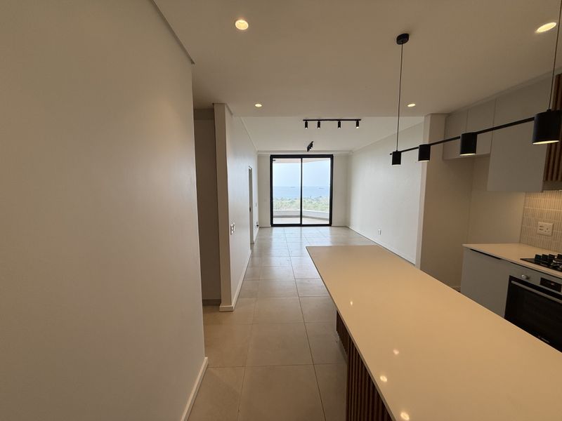 Modern open-plan living area featuring a sleek kitchen island, built-in appliances, tiled floors, and large sliding doors leading to a balcony with ocean views.