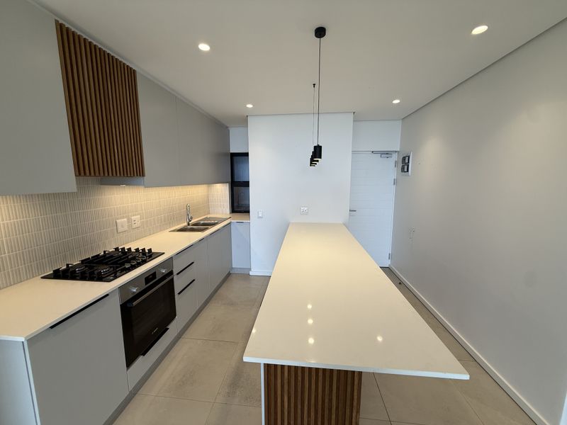 Modern kitchen featuring a long island, gas hob, built-in oven, sleek cabinetry, and contemporary lighting in an open-plan layout.