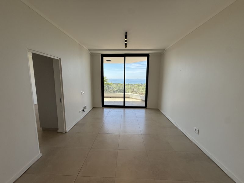 A bright, empty living area featuring large sliding glass doors that open to a balcony with scenic ocean views and modern tiled flooring.