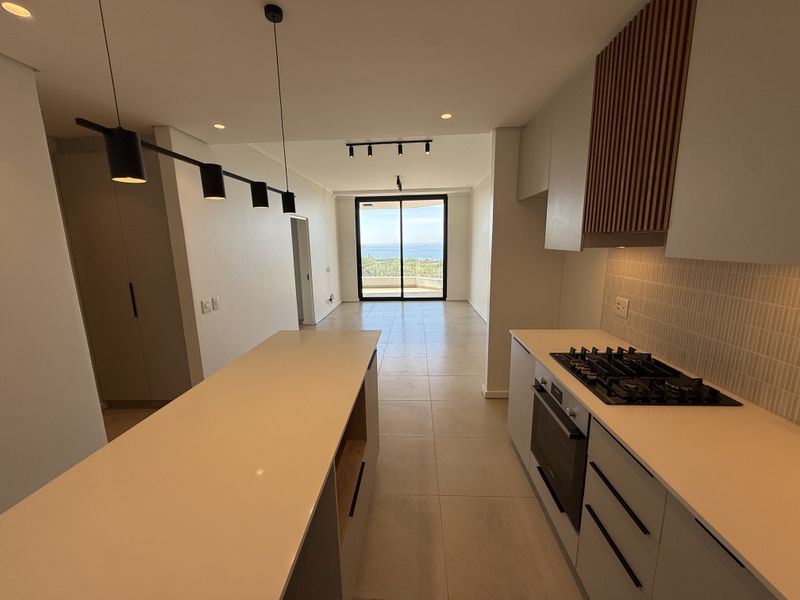 Modern open-plan kitchen and living area featuring a central island, gas cooktop, sleek cabinetry, and large sliding doors leading to a balcony with ocean views.