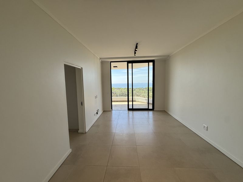 An empty, well-lit living area featuring large format floor tiles and a sliding glass door leading to a balcony with scenic views.
