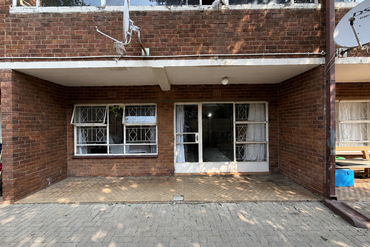 Rear exterior view of an apartment featuring exposed brickwork, a tiled patio area, paved ground, and security-fitted windows and sliding doors.