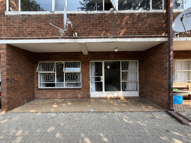 Rear exterior view of an apartment featuring exposed brickwork, a tiled patio area, paved ground, and security-fitted windows and sliding doors.