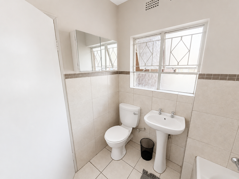 A clean, functional bathroom featuring a pedestal basin, toilet, tiled finishes, and a mirrored cabinet with natural light from a window with security bars.