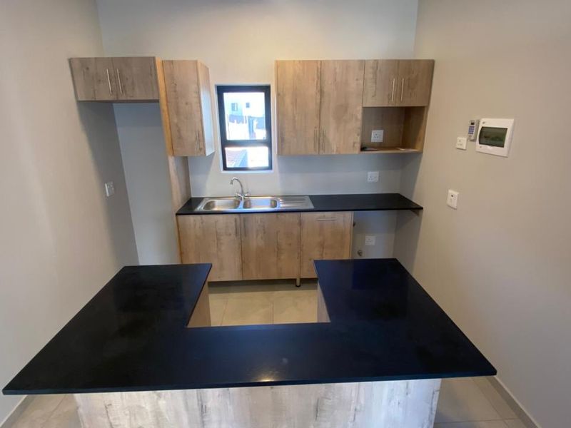 A modern, empty kitchen featuring light wood-finish cabinetry, dark countertops, a breakfast bar, and a stainless steel sink.