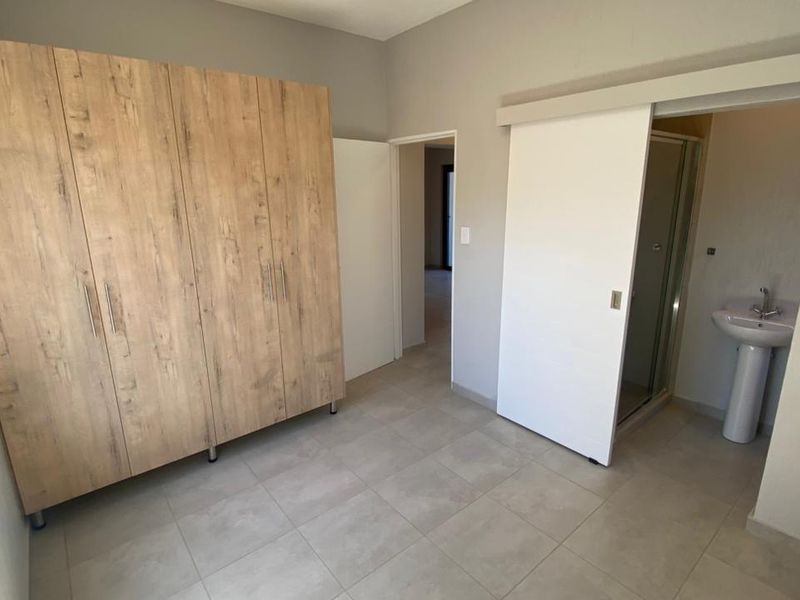 A clean, modern bedroom featuring built-in wooden cupboards, tiled flooring, and an en-suite bathroom with a shower and pedestal basin.