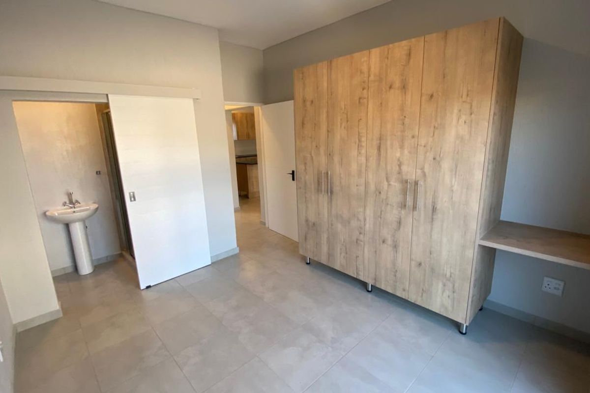 A spacious bedroom featuring neutral tiled flooring, a large built-in wardrobe with an integrated desk, and an en-suite bathroom with a pedestal basin.