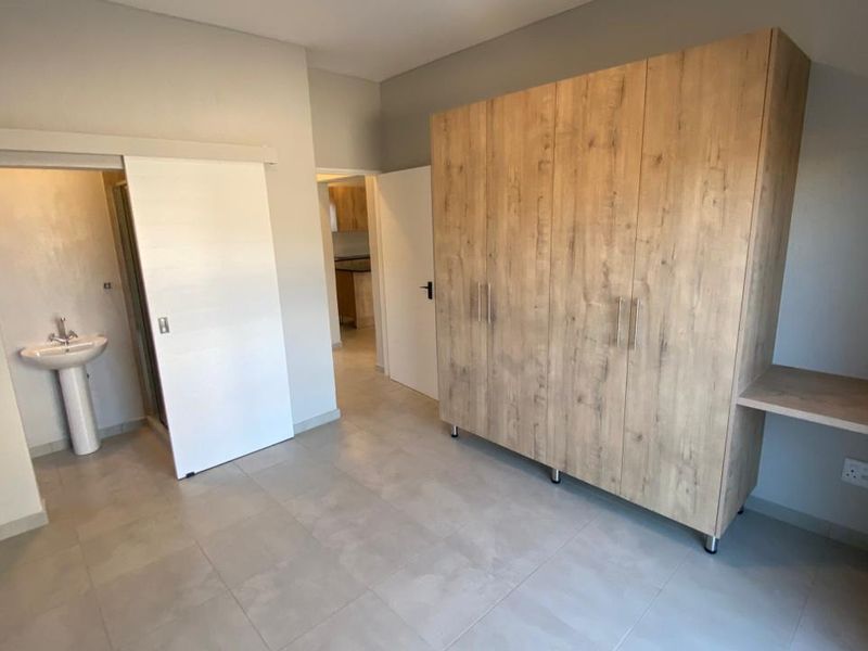 A spacious bedroom featuring neutral tiled flooring, a large built-in wardrobe with an integrated desk, and an en-suite bathroom with a pedestal basin.