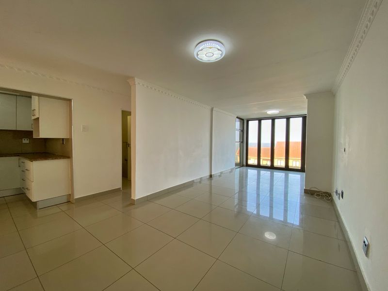 Spacious open-plan living area featuring tiled floors, ceiling cornices, and a compact kitchen, leading to a balcony with natural light.