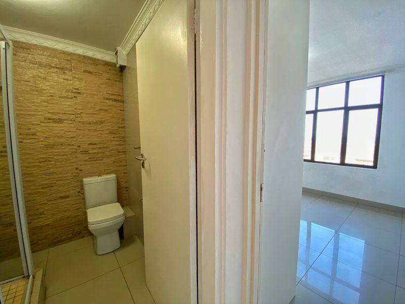 A clean, functional bathroom featuring a toilet, shower enclosure, and textured wall tiling, adjacent to a bright, tiled living area.