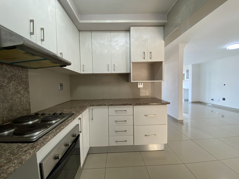 A clean, modern kitchen featuring white cabinetry, laminate countertops, built-in oven and hob, and neutral tiled flooring, flowing into an open-plan living area.