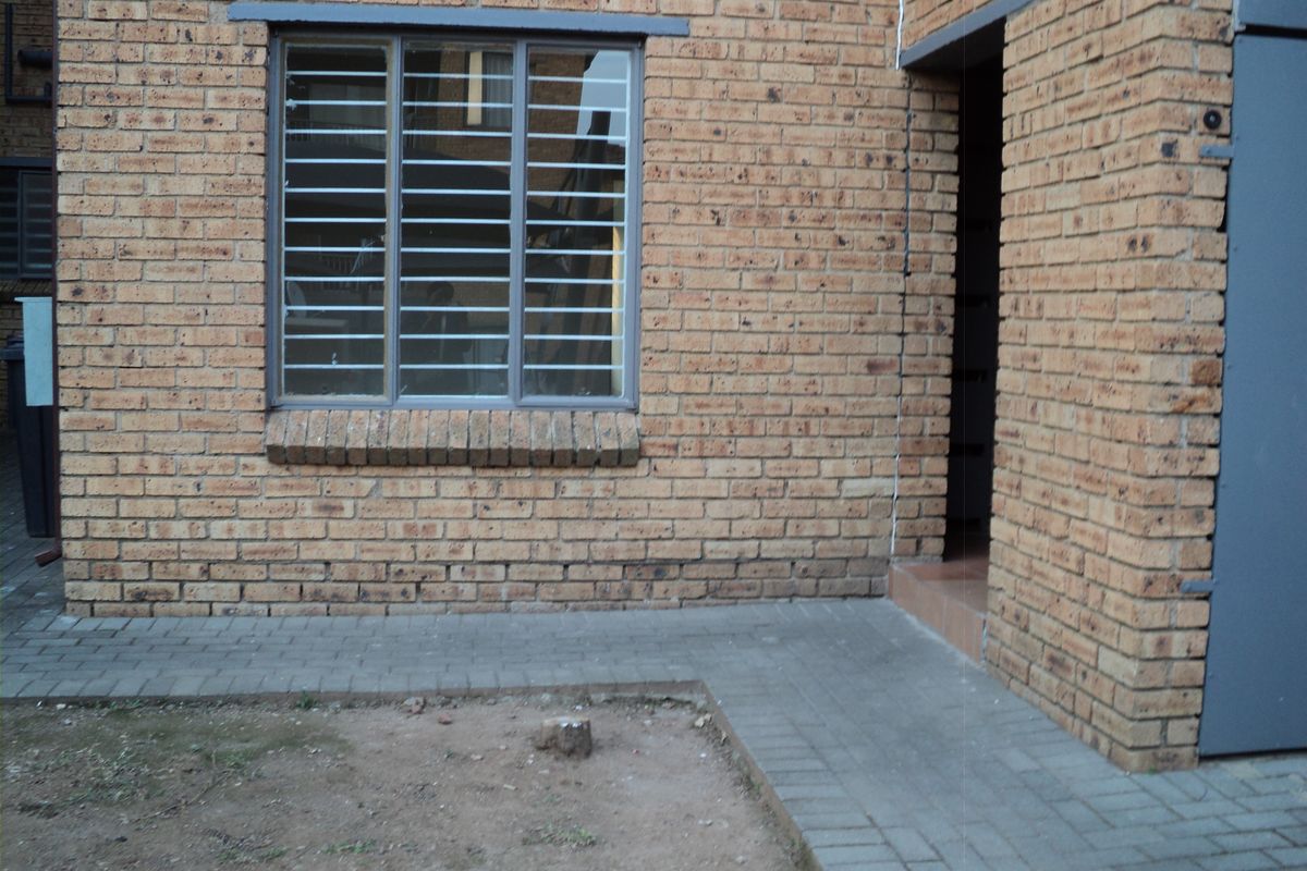 Rear exterior view of the apartment featuring a face brick facade, security-barred window, and a paved walkway leading to the entrance.